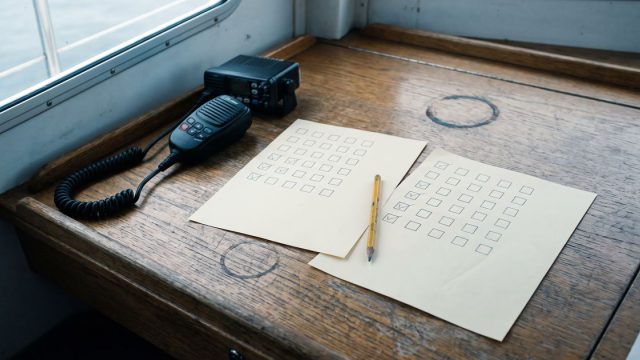 Ein UKW-Funkgerät auf einem hölzernen Kartentisch, daneben zwei Antwortbögen mit Kästchenrastern — die angekreuzten Kästchen sitzen auf beiden Bögen an verschiedenen Stellen.