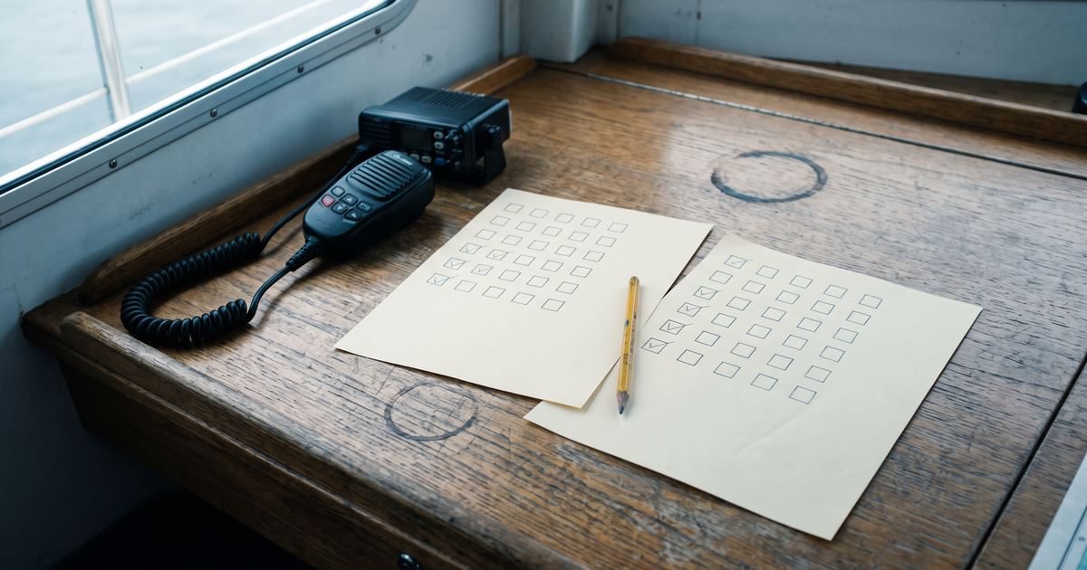 Ein UKW-Funkgerät auf einem hölzernen Kartentisch, daneben zwei Antwortbögen mit Kästchenrastern — die angekreuzten Kästchen sitzen auf beiden Bögen an verschiedenen Stellen.