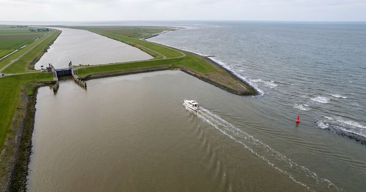 Luftaufnahme einer Flussmündung: links Binnenwasser zwischen Deichen mit einer Schleuse, rechts offene See mit einer roten Tonne, dazwischen ein weißes Motorboot.
