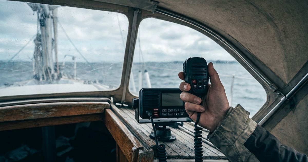 Hand hält das Hörerteil eines schwarzen UKW-Seefunkgeräts im Cockpit eines Segelboots, im Hintergrund graues Wasser unter Wolken.